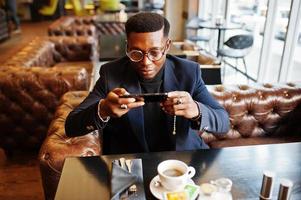 Fashionable african american man in suit and glasses sitting at cafe and snap coffee on phone. photo