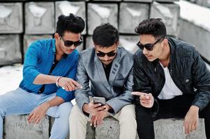 grupo de tres jóvenes indios casuales con gafas de sol posados contra bloques de piedra y mirando el teléfono móvil. foto