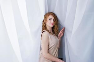 Curly hair girl on beige dress posed at white curtains. photo
