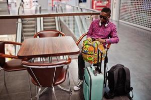 African american man in checkered shirt, sunglasses and jeans with suitcase and backpack. Black man traveler on duty free. photo