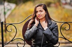 Pretty indian girl in black saree dress and leather jacket posed outdoor at autumn street and sitting on bench. photo