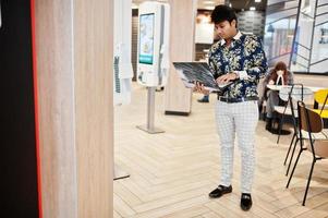 joven autónomo indio sentado con una computadora portátil abierta en un café de comida rápida, un apuesto hombre asiático leyendo un mensaje de texto durante el trabajo en un net-book en una cómoda cafetería. foto