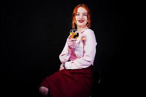 Gorgeous red haired girl in pink blouse and red skirt with glass of wine at hand isolated on black. photo