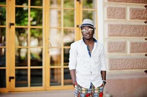 Stylish african american man in white shirt and colored pants with hat and glasses posed outdoor. Black fashionable model boy. photo