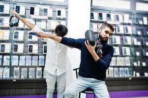 Two indians mans customer buyer at mobile phone store with earphones and wireless speaker listening music and throws dab. South asian peoples and technologies concept. Cellphone shop. photo