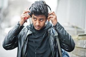 Stylish and casual asian man in black leather jacket, headphones with red mobile phone at hands posed on street and listening music. photo
