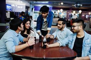 Group of stylish asian friends wear on jeans sitting at table and drink soda from bottles in club. photo