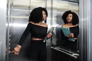 Fashionable young beautiful african american business woman with afro hairstyle wear in elegant black, stand at elevator with phone and laptop at hands. photo