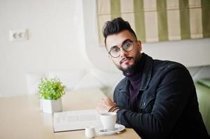 Arab man wear on black jeans jacket and eyeglasses sitting in cafe, read book and drink coffee. Stylish and fashionable arabian model guy. photo