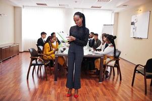 Face of handsome african business woman, holding clipboard on the background of business peoples multiracial team meeting, sitting in office table. photo