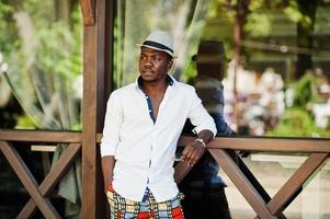 elegante hombre afroamericano con camisa blanca y pantalones de colores con sombrero al aire libre. chico modelo de moda negro. foto