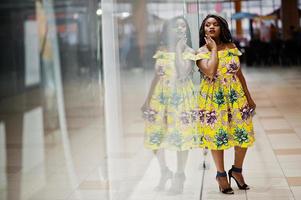 Cute small height african american girl with dreadlocks, wear at coloured yellow dress, posed of shop showcase at trade center. photo