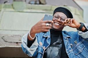 African american man in jeans jacket, beret and eyeglasses, with cigar posed against btr military armored vehicle, and making selfie on mobile phone. photo