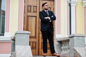 Stylish indian businessman in formal wear standing against door in business center with cross arms. photo