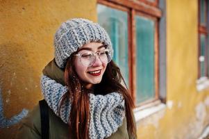 Portrait of brunette girl in gray scarf and hat, glasses at cold weather with sunshine against orange wall of old house. photo