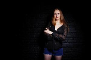 Studio shot of brunette girl in black blouse with bra and shorts against black brick wall. photo