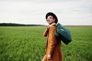 Stylish man in glasses, brown jacket and hat with bag posed on green field. photo