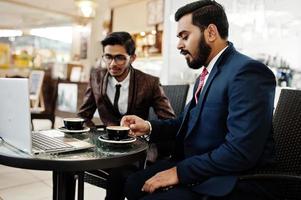 Two indian business man in suits sitting at office on cafe, looking at laptop and drinking coffee. photo