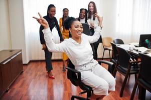 Five multiracial business womans standing at office and roll woman on chair. Diverse group of female employees in formal wear having fun. photo