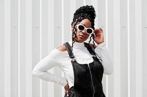 African woman in denim overalls skirt, white sunglasses posed against white steel wall. photo