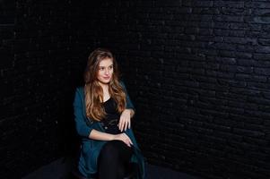 hermosa chica morena vestida con una chaqueta negra y verde, sentada y posando en una silla en el estudio contra una pared de ladrillo oscuro. retrato de modelo de estudio. foto