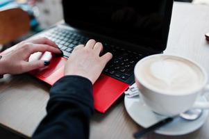 cierra las manos de una chica que trabaja con un portátil rojo con una taza de capuchino. foto