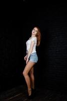 Portrait of a gorgeous redheaded girl in white t-shirt and denim shorts posing in the studio next to the brick wall. photo