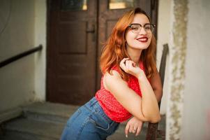 atractiva mujer pelirroja con anteojos, vestida con blusa roja y falda de jeans posando. foto