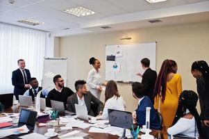 Mixed race business coach presenting report standing near whiteboard pointing on sales statistic shown on diagram and chart teach diverse company members gathered together in conference room. photo