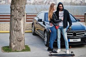 Cool multiracial couple hugs together with longboard against black elegant sport car. photo