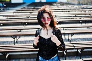Portrait of brunette girl in pink glasses wear on black posed outdoor on sunny day against row of benches. photo