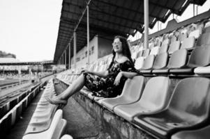 Portrait of a young beutiful girl in dress and sunglasses sitting on the tribunes in stadium. photo