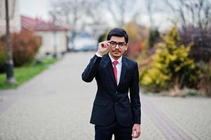 joven indio con anteojos, traje negro con corbata roja posado al aire libre. foto