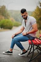 el hombre de barba árabe alto de moda usa camisa, jeans y gafas de sol sentado en un banco rojo en el parque. foto