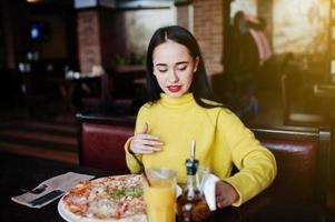 Funny brunette girl in yellow sweater eating pizza at restaurant. photo