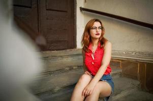 Attractive redhaired woman in eyeglasses, wear on red blouse and jeans skirt posing. photo