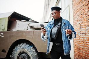 African american man in jeans jacket, beret and eyeglasses, smoking cigar and posed against btr military armored vehicle, with mobile phone at hand. photo