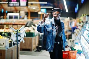 elegante hombre afroamericano casual con chaqueta de jeans y boina negra sosteniendo una canasta y leche a mano, parado cerca de la nevera y comprando en el supermercado. foto