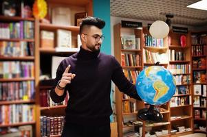 estudiante árabe alto e inteligente, vestido con cuello alto violeta y anteojos, en la biblioteca sosteniendo el globo terráqueo en las manos. foto