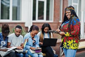 Group of five african college students spending time together on campus at university yard. Black afro friends studying at bench with school items, laptops notebooks. photo