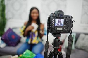 Cute african american woman making a video for her blog using a tripod mounted digital camera. Young female blogger or vlogger on camera. photo