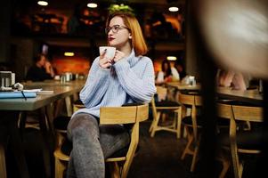 Cheerful young beautiful redhaired woman in glasses sitting at her working place on cafe and drinking coffee. photo