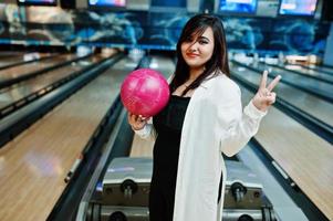 elegante mujer asiática de pie en la bolera con la pelota en la mano y muestra dos dedos. foto