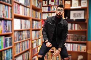 estudiante árabe alto e inteligente, vestido con chaqueta negra de jeans y anteojos, en la biblioteca sentado en una silla contra estantes de libros. foto