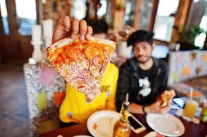 amigos asiáticos chicos comiendo pizza durante la fiesta en la pizzería. gente india feliz divirtiéndose juntos, comiendo comida italiana y sentados en el sofá. foto