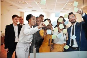 Team of multicultural young people pointing on glass with colorful paper notes. Diverse group of male and female employees in formal wear using stickers. photo