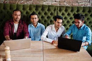 grupo de cuatro hombres del sur de asia posaron en una reunión de negocios en un café. los indios trabajan juntos con computadoras portátiles usando varios dispositivos, conversando. foto