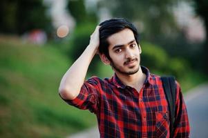 joven estudiante indio con camisa a cuadros roja y jeans con mochila posada en la calle. foto