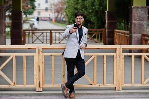 Stylish indian business man with bindi on forehead and glasses, wear on gray suit posed outdoor and looking at mobile phone. photo