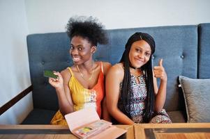 Two black african girlfriends at summer dresses sitting in cafe with credit card at hand. photo